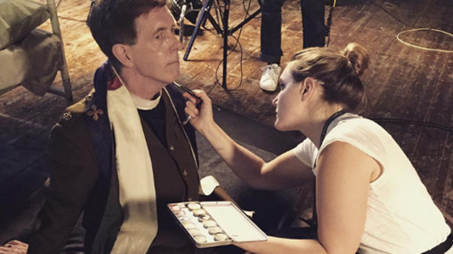 Make-up artist applying colour to an actor in full costume on the set of a TV programme